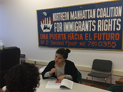 Woman sitting at a table teaching other people and behind her is the sign for the Northern Manhattan Coalition for Immigrants Rights.